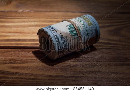 A Roll Of Dollars Lies On A Wooden Textured Table In The Dark Illuminated By A Ray Of Light.