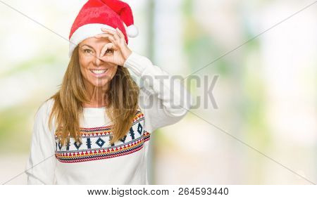 Middle age adult woman wearing winter sweater and chrismat hat over isolated background doing ok gesture with hand smiling, eye looking through fingers with happy face.