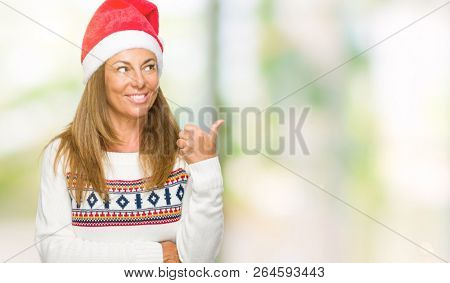 Middle age adult woman wearing winter sweater and chrismat hat over isolated background smiling with happy face looking and pointing to the side with thumb up.