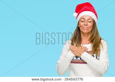Middle age adult woman wearing winter sweater and chrismat hat over isolated background smiling with hands on chest with closed eyes and grateful gesture on face. Health concept.