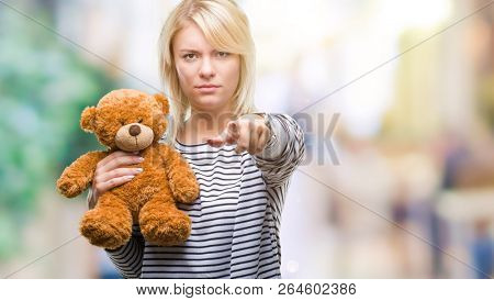 Young beautiful blonde woman holding teddy bear plush over isolated background pointing with finger to the camera and to you, hand sign, positive and confident gesture from the front