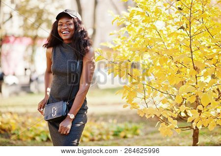 Stylish African American Girl In Gray Tunic, Crossbody Bag And Cap Posed At Sunny Autumn Day Against
