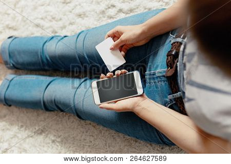 Young Woman Holding Credit Card And Using Phone Sitting On Floor. Payment.  Online Shopping Concept.