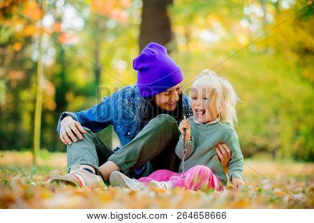 Portrait Of A Mother With A Child Girl Sitting On A Ground In A Prak. Fall Season Time
