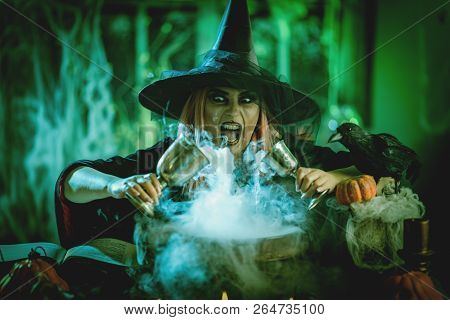 A Witch In Foggy Surrounding Holds Two Goblets Above Boiling Water For A Cooking Poison Soup.