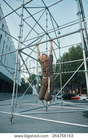 Portrait Of A Happy Young Woman Having Good Time While Hanging On Metallic Construction Outdoors. Ur