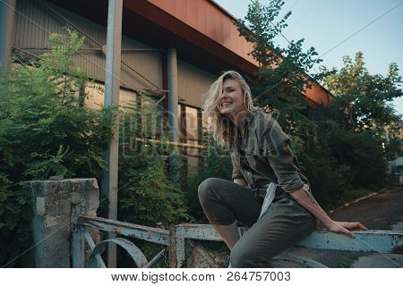 Stylish Girl Posing In The Street Sitting On A Steel Fence With Bent Legs. Urban Style. Street Fashi