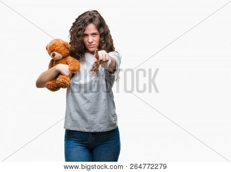 Young brunette girl holding teddy bear over isolated background pointing with finger to the camera and to you, hand sign, positive and confident gesture from the front