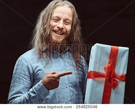 Happy Male With Long Hair And Beard In Blue Shirt Pointing On Gift Box In His Hand. Christmas Concep