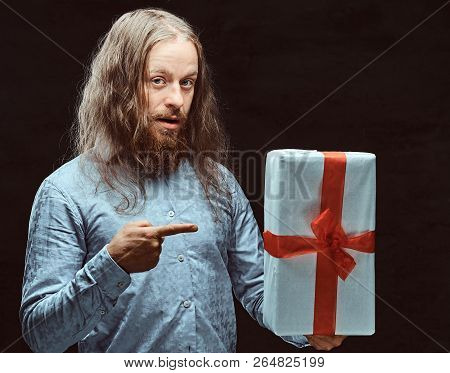 Happy Male With Long Hair And Beard In Blue Shirt Pointing On Gift Box In His Hand. Christmas Concep