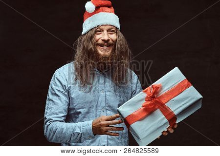 Happy Male With Long Hair And Beard In Blue Shirt And Red Santa Hat Holds Gift Box.