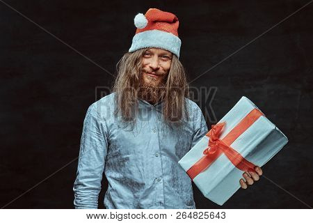 Happy Male With Long Hair And Beard In Blue Shirt And Red Santa Hat Holds Gift Box.