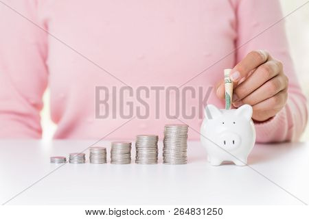 Woman Hand Putting America Dollars Banknotes Money Into Piggy Bank With Stack Of Coins And Alarm Clo