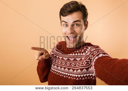 Image of young man 20s with bristle wearing knitted sweater pointing finger aside while taking selfie photo isolated over beige background