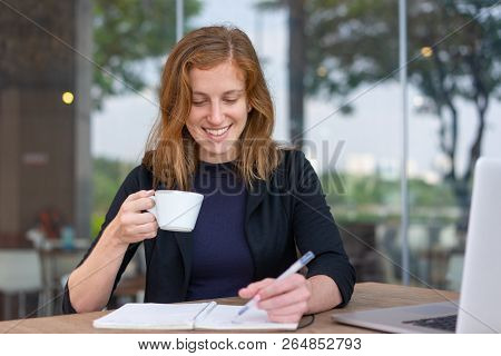 Positive Student Reading Copybook During Coffee Break. Young Woman In Dark Blue Jacket Drinking Coff