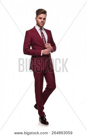 elegant young man in grena suit stepping on white background while fixing cuffs