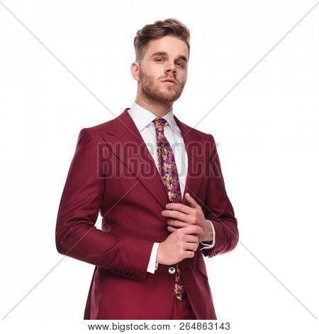portrait of proud man iwearing an elegant grena suit arranging his ring while standing on white background