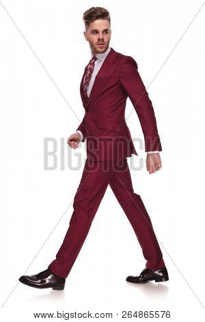 side view of confident businessman in grena suit walking on white background and looking behind, full body picture