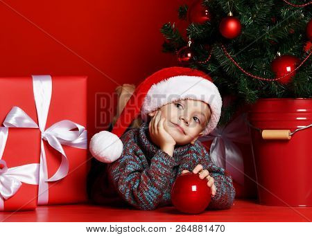 Funny Smiling Childboy In Santa Red Hat Lying On  Christmas Tree  Holds In One Hand A Christmas Ball