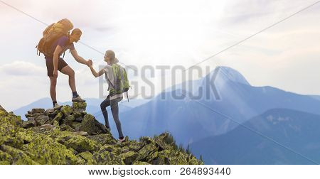 Young Tourists With Backpacks, Athletic Boy Helps Slim Girl To Clime Rocky Mountain Top Against Brig