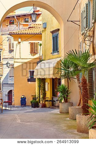 Lovran, Croatia. Cosy streets of ancient old town with arc, street lantern, vintage buildings and palm trees in buckets. Sunny summer day. Urban landscape of historical centre.