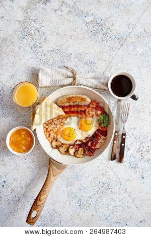 Full English Breakfast served in a pan. Fried eggs, beked beans, tomatoes, champignons, crispy bacon, sausages and toast. Placed on stone background. Top view with copy space.