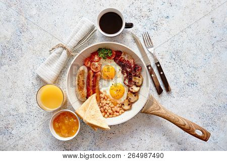 Full English Breakfast served in a pan. Fried eggs, beked beans, tomatoes, champignons, crispy bacon, sausages and toast. Placed on stone background. Top view with copy space.
