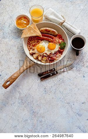 Full English Breakfast served in a pan. Fried eggs, beked beans, tomatoes, champignons, crispy bacon, sausages and toast. Placed on stone background. Top view with copy space.