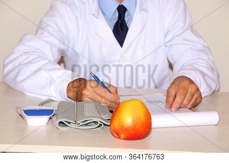Close-up - The Doctor In A White Coat Sits At The Table And Eats A Healthy Apple.