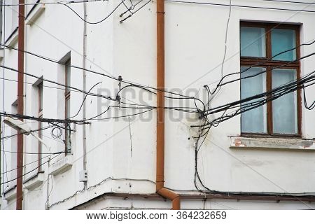 The Plastered Facade Of An Old House. Plenty Of Entangled Electrical Wires Near Windows And A Drainp