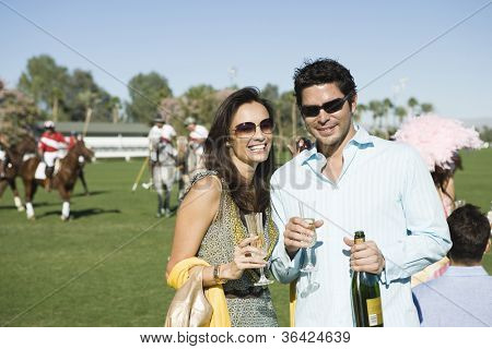 Ritratto di coppia felice avendo champagne con i giocatori giocare a polo in background