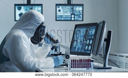 Scientist in protection suit and masks working in research lab using laboratory equipment: microscopes, test tubes. Coronavirus 2019-ncov hazard, pharmaceutical discovery, bacteriology and virology.