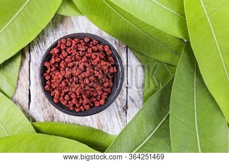 Bowl With Red Annatto Grains Surrounded By Green Leaves On Wooden Background, Top View