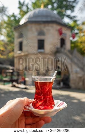 Turkish tea in a traditional turkish garden in Koza Han Silk Bazaar in Bursa, Turkey. Koza Han is historical place from Ottoman. Hand holding glass of tea with Koza Han at background