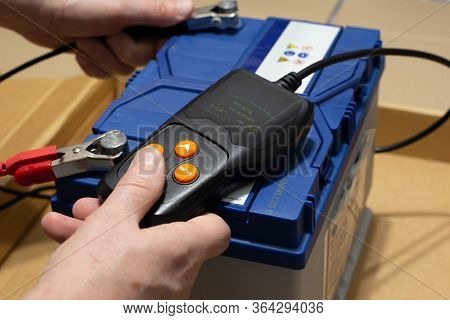 Man Carries Out Diagnostics Of A Car Battery With A Tester, Closeup. Akb Good-translation