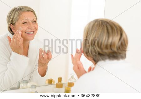 Starší žena v zrcadle a použitím anti-wrinkles krém