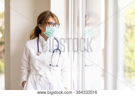Female Doctor Wearing Face Mask And Looking Careworn While Looking Out The Window During Coronavirus