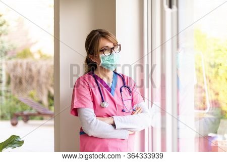 Female Nurse Wearing Face Mask And Looking Careworn While Looking Out The Window During Coronavirus 