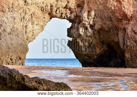 Empty Beach Between Cliffs And Natural Archway Through Which You Can See The Sea. Uncrowded Travel C