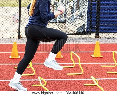 Side View Of High School Girl Stepping Over Yellow Mini Hurdles During Speed Drills At Track And Fie
