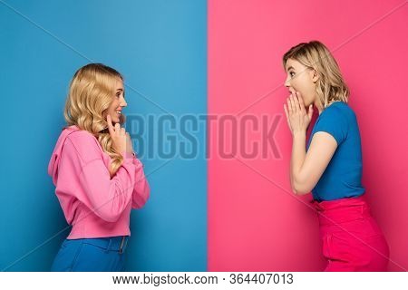 Side View Of Shocked And Smiling Blonde Sisters Looking At Each Other On Pink And Blue Background