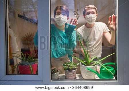 Man And Woman With Medical Mask On Their Faces Looking Through Window, Stay At Home, Selfisolation T