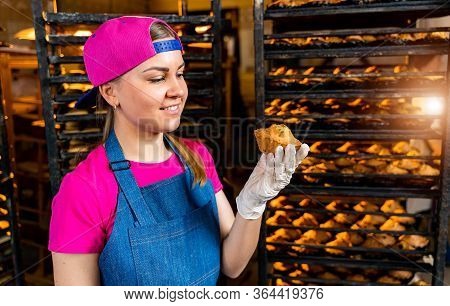 Young Happy Female Worker In Sterile Cloths Holding Freshly Baked Muffins On Tinplate Inside Food Pr