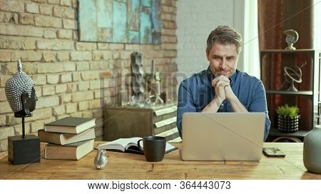 Older man tele working with laptop computer at home office in living room, thinking, looking busy.
