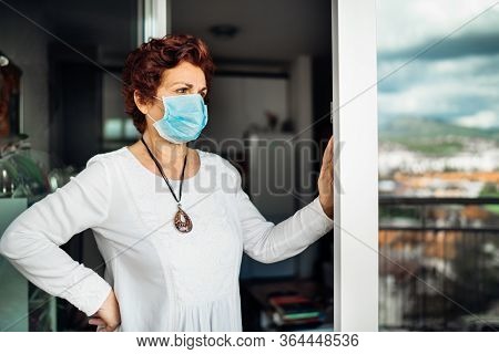 Senior Elderly Sad Woman At Home,wearing Mask On Balcony Window.coronavirus Covid-19 Disease Outbrea