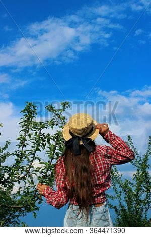 Young  Brunette Girl Wearing A Summer Hat, Red Shirt And Blue Jeans Satnding Near Green Trees And Lo