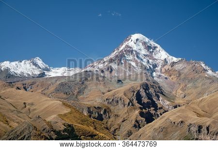 Snow Covered Mountain Kazbeg With Gergeti Trinity Church In The Foreground With Blue Sky Above, Step
