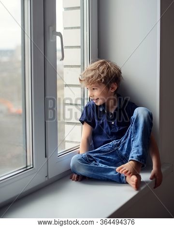 Little Cute Boy Sits On A Window Sill, Looks Out The Window And Smiles.