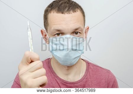 Man In A Medical Mask Holds A Thermometer In His Hand On A Light Background