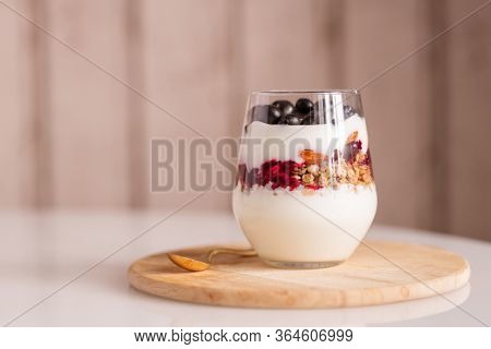 Glass with several layers of fresh yoghurt ingredients - sourcream, cherry jam, muesli and almond kernels - standing on wooden board on table